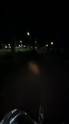 The video captures a first-person perspective of someone riding a bicycle at night on a paved path. The path is lined with trees and intermittent streetlights illuminate the surroundings. The light from the bicycle's front lamp casts a dim, warm glow on the path directly ahead. In the distance, more streetlights are visible, creating a series of faint lights along the path. The overall atmosphere is dark and quiet, with the focus being on the illuminated path and the surrounding dimly lit environment.