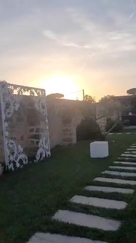 The video showcases a serene outdoor garden and pool area during the golden hour, likely sunrise or sunset, in Sogliano Cavour, Italy. The scene opens with a view across a lush green lawn, where a stone-slab pathway leads towards a modern outdoor covered structure featuring stone pillars and a dark roof. A white cube-shaped piece of outdoor furniture rests on the grass. On the left, traditional stone walls and trees are visible, bathed in the warm, low-angle light.

As the camera pans to the right, it reveals a spacious, rectangular swimming pool filled with clear blue water. The pool's surface beautifully reflects the sky and the surrounding structures, including a white metal pergola over parts of the garden. The pool is edged with light-colored coping, and two circular lights or features are visible at its bottom. The background features more green landscaping, stone walls, and scattered trees. The sky is bright with some faint clouds, indicating pleasant weather. No people or visible text are present, emphasizing the tranquil and inviting atmosphere of this well-maintained outdoor space.