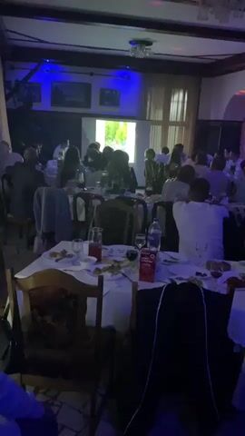 This is a video taken inside a dimly lit restaurant or event hall. The scene is filled with people seated at round tables covered with white tablecloths. The tables are laden with food and drinks, suggesting a banquet or celebratory gathering. The lighting is primarily blue and purple, creating a festive and somewhat dramatic atmosphere. In the background, a projector screen displays an image, and there's a stage area where a few individuals appear to be performing or presenting. Some people are actively engaged, while others seem to be observing. The overall mood appears to be one of conviviality and entertainment. There are no explicit weather cues visible as the scene is indoors. The time of day is not specified, but the indoor setting and lighting suggest it could be evening or night. There is no visible text in the frame.