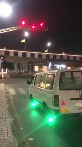 At night, a white van with green underglow lights is parked at a crosswalk with a red traffic light overhead. The van is facing away from the camera, its rear is visible. A yellow and black toll booth-like structure with lit-up stripes and flags stands behind the van. A street lamp illuminates the scene from the left. The crosswalk is striped white on the asphalt. There are buildings visible in the distance beyond the toll booth structure. A sign on the left of the frame has a phone number displayed.