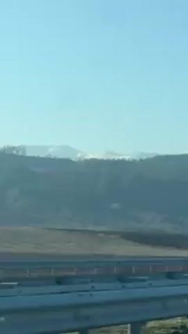 The video shows a landscape seen from a moving vehicle, likely a car, traveling on a highway. A gray guardrail is visible in the foreground, along with a field and some low-lying vegetation in the middle ground. Beyond this, there is a forested area with hills, and in the far distance, there are snow-capped mountains under a clear blue sky.  The overall impression is one of a sunny, peaceful drive through a rural area.  A highway sign is visible near the middle of the video.