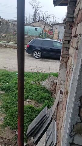 The video shows a black Audi A4 Avant parked on a dirt road in a residential area. The car is seen from behind a brick wall, with a metal pole partially obstructing the view. There is green vegetation and some scattered debris near the wall. Other houses and structures are visible in the background. The sky is overcast.