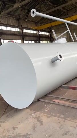 A large, grey, cylindrical tank is shown lying on its side inside a warehouse. The tank has several flanges attached to its exterior, and a pipe extends horizontally above it. The warehouse has a concrete floor, exposed metal beams, and windows in the ceiling. There are no people visible in the video. The camera pans slowly along the length of the tank.
