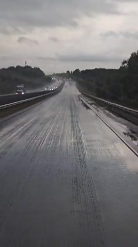 This video captures a drive on a wet highway in Vislovka, Russia. The scene is set on a multi-lane road with trees lining both sides. The sky is overcast with dark clouds, suggesting it is either early morning or late evening, or simply a very cloudy day. The road surface is slick with water, indicating it is raining or has recently rained heavily. Water splashes are visible on the road, and the lanes are marked with white lines. Several vehicles are present, including cars and trucks, navigating the wet conditions. One white car is particularly prominent in the foreground, its taillights visible as it drives away. There is no clear indication of the specific time of day beyond the overcast sky. The mood conveyed is one of a typical drive, possibly with a sense of caution due to the weather. A road sign with the number "1077" is visible on the left side of the highway. The overall impression is a somewhat dreary but functional highway scene in Russia.