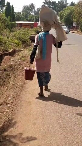A person walks along a paved road in what appears to be a rural or semi-rural setting. They are seen from the back, carrying a large sack on their head, a bucket in their left hand, and a small bag slung over their shoulder. The person is wearing a light-colored shirt, a dark skirt with stripes, and sandals. They also have a light blue scarf around their neck. The road is flanked by grassy areas and trees. A building is visible in the background, along with a passing vehicle.  The weather seems sunny. The person's shadow is cast on the road.