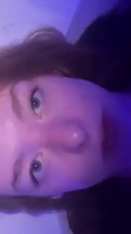The video shows a person lying in a tub of water, immersing their face and hair. The person is light-skinned with red hair and fair-colored eyes. Their face is wet and has visible freckles or spots on the nose. They are wearing lip gloss or lipstick. Water flows over the person’s face and hair as they submerge themselves and come back up. The background is white, possibly the interior of a bathtub. The lighting is somewhat dim, contributing to a soft, almost ethereal quality of the image.