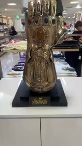 The video shows a gold-colored Infinity Gauntlet replica on a black base with the Avengers Infinity War logo. A hand with a white ring and a metal wristwatch is placed next to the replica for scale. The gauntlet is displayed inside a store with clothing items visible in the background and people browsing.