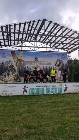 A group of people stand on a stage in front of a large banner. The banner features images of film strips and trees, with text in Russian that appears to say "Tourist Camp." Seven men are gathered in the center of the stage, most of whom are wearing dark clothing. One man on the left is playing a guitar. To the right of the group of men, a man in a bright yellow safety vest stands next to two women in white shirts. The stage is covered by a large, transparent roof supported by metal beams. The background shows a cloudy sky and trees. The stage is set up outdoors, with a grassy area in the foreground.