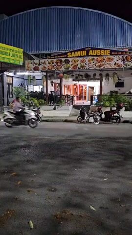 The video shows a nighttime street scene in front of a restaurant called Samui Aussie. The restaurant has a large sign with pictures of food. There are tables and chairs outside the restaurant. Several motorcycles are parked on the side of the road, and others are driving by. One motorcycle in the foreground is blurred due to motion. The road is wet and reflective. There are some fallen leaves on the ground.