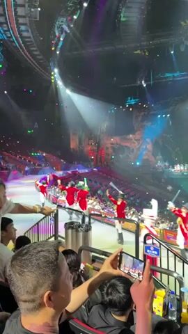 The video shows a view from a theater audience watching a show. The stage is well-lit with colorful lights, with performers dressed in red coats and black hats moving across the stage with swords. There is a large white screen in the center of the stage, along with elaborate set designs. The audience members are seated in tiers, with some people recording the performance on their phones. A metal railing separates the audience from the level below. The theater appears large and crowded.