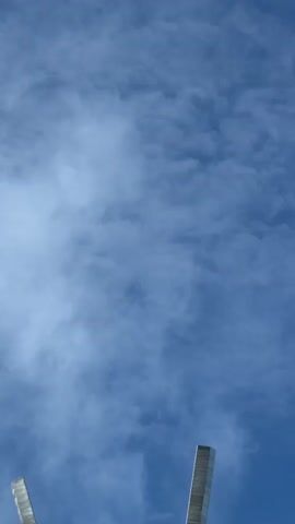 The video shows a low-angle shot of a sculpture against a blue sky with clouds. The sculpture appears to be made of metal and consists of several tall, thin, rectangular elements radiating upwards from a central point. There is also a circular metal element visible at the base of these radiating pieces. The camera slowly zooms out, revealing more of the sculpture and the surrounding sky.