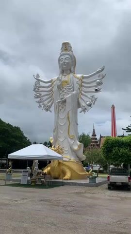 A large, multi-armed statue of a deity, likely Guanyin, stands prominently in an outdoor setting. The statue is predominantly white with gold accents and rests on a golden, reptilian-like base. Numerous smaller statues and offerings are placed in front of the large statue, some under a white tent. In the background, there are trees, several structures with traditional Asian architectural elements, and a tall, red and white striped tower or chimney. A light-colored pickup truck is parked to the right of the frame. The sky is overcast. The video appears to be a static shot or has minimal camera movement.