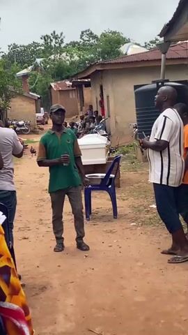 The video shows an outdoor scene in what appears to be a village setting. In the foreground, a person wearing a colorful patterned garment is partially visible on the left. In the center of the frame, a man wearing a green shirt with brown sleeves and a cap, holding a bottle, stands facing the camera. To his right, a white coffin is visible, along with a blue plastic chair. Further to the right, a man in a white and black striped shirt and dark pants is engaged in conversation with someone off-camera. Behind them, several people are gathered, and houses with corrugated metal roofs and brick walls are visible. Motorcycles are parked along the path, and the ground is dirt. The overall atmosphere suggests a gathering, possibly a funeral or a community event.