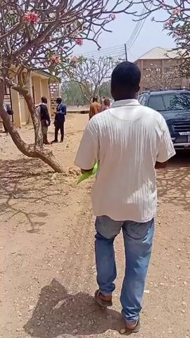 The video shows a man walking away from the camera on a dirt road. The man is wearing a white shirt and blue jeans. He is carrying a green folder in his right hand. There are several other people standing in the distance, some of whom are talking. There is a car parked on the right side of the road, and a tree with pink flowers on the left side. The ground is dry and there are small bits of debris scattered around.
