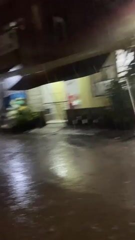 The video shows a dark, wet street at night, likely due to rain. Reflections of streetlights and building lights shimmer on the slick surface of the road. Buildings line the side of the street, with some appearing to be businesses, as indicated by signs and awnings. There are hints of people or vehicles in the distance, blurred by the motion of the camera and the low light conditions. The overall atmosphere is moody and atmospheric, suggesting a city street during a rainy night.