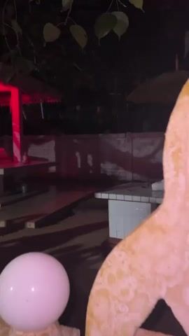 The video shows a small, illuminated structure, possibly a shrine or altar, at night. The structure is lit with bright red lights, creating a dramatic and somewhat eerie ambiance. The building itself is white with decorative trim. In front of the structure, there are wooden planks laid out, perhaps to create a temporary path or seating area. The ground appears to be paved or made of concrete. Dark foliage hangs from the top of the frame, suggesting the presence of trees or other plants. The overall scene is dimly lit, with the red glow of the structure being the primary source of light. There are no visible people in the frame. The video is shot from a low angle, with a large, blurred object in the foreground, possibly part of the camera or another object obscuring the lower left portion of the scene.