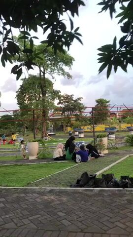 The video shows a group of six young people sitting on the grass in an outdoor area. The group is facing away from the camera, and it is unclear what they are doing. The environment appears to be a park or a recreational area, with trees, grass, and paved walkways. There are also some planters and decorative elements visible in the background. The sky is cloudy and the light suggests it is either early morning or late afternoon. One person is wearing a blue shirt, another a green shirt, and two are wearing hijabs. The overall impression is one of a casual gathering in a pleasant outdoor setting.