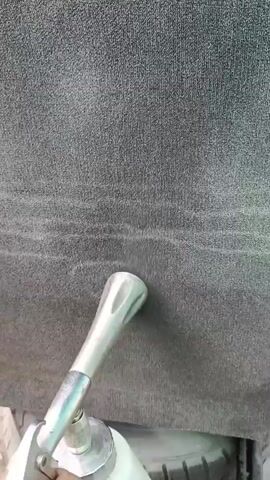 A hand holds a foam gun and sprays foam onto a grey fabric surface, possibly upholstery or carpet. The foam gun has a metallic nozzle and a white plastic bottle. The fabric appears to be textured and shows some lighter lines where the foam is applied. In the background, a tire is visible, suggesting the location may be a garage or workshop. The video is shot from a first-person perspective.