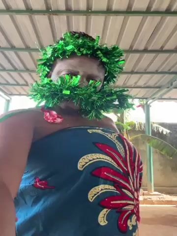 A dark-skinned woman, wearing a blue and red patterned wrap around her torso and a green tinsel garland partially obscuring her face, is standing under a covered structure. The structure has a corrugated metal roof held up by a green metal frame. Behind the woman, a concrete wall is visible, along with some banana trees. The ground appears to be dirt or sand. The woman is moving her arms and body slightly.