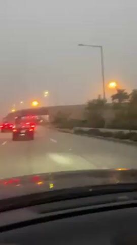 The video shows a car's dashboard view of a road with heavy fog, featuring streetlights and other cars. The road has multiple lanes and a median with palm trees. A McDonald's restaurant sign is visible on the left side of the road. Other vehicles, mostly with red taillights, are visible ahead, indicating traffic. The camera is inside the car and shows part of the windshield and dashboard, which appear to be wet with water droplets, suggesting it might be raining or drizzling. The overall visibility is poor due to the dense fog, which obscures the background and makes the scene look muted.