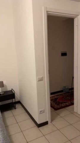 The camera pans around a bedroom in an Airbnb in Italy. The walls are white and the floor is tiled. There is a double bed with a gray headboard and two decorative pillows. Above the bed hangs a large painting depicting what appears to be an angelic scene. On either side of the bed is a small black nightstand, each with a lamp on it. There is a doorway leading to another room with a red rug. There is an air conditioning unit on the wall.