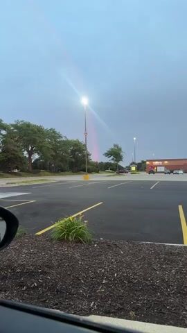A security camera captures a parking lot with a light pole in the center. The sky is cloudy and overcast. There is a faint rainbow visible to the right of the light pole. The parking lot is empty except for a few cars parked in the distance. There are trees on the left side of the frame and a building on the right. The camera is positioned low to the ground, giving a unique perspective of the scene. There is a dark car in the foreground on the left.