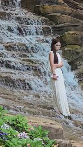 A young woman in a white outfit stands on a rock next to a multi-tiered waterfall. She is holding a phone in her left hand and her right arm is bent across her chest. The background is a rocky waterfall with water flowing down it and some green foliage in the foreground.