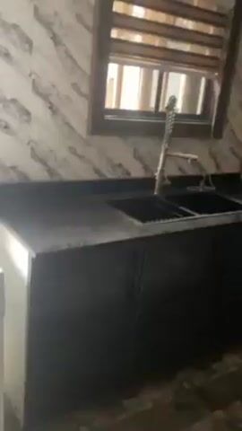 The video presents a view of a kitchen. The kitchen features dark gray cabinetry with dark handles and a black countertop. There is a stainless steel sink with a faucet, and above the sink, a window with blinds. The walls are decorated with wallpaper that has a marbled pattern in shades of white and gray. Above the cabinets on the opposite side of the sink are red cabinets and some black shelving. Built into the cabinets are a microwave oven. A cardboard box is visible on the counter, in the corner. The lighting is ambient, giving the room a somewhat subdued ambiance.  The video appears to be a tour of the kitchen, as the camera pans slowly to the right to show the extent of the cabinetry.