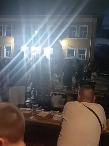 The video shows an outdoor gathering of people in front of a building. Bright lights illuminate the scene, creating glare. In the foreground, a person with a shaved head is visible from the back, sitting at a wooden table. On the table are various items, including a plate with food, glasses, and bottles. Several other people are standing and sitting around the table and further back, in front of the building. Some individuals appear to be interacting. A green umbrella is visible behind the crowd. The building has windows and an entrance area that is brightly lit, suggesting an event or activity is taking place inside or just outside. The time of day appears to be evening or night, given the dark sky in the background.
