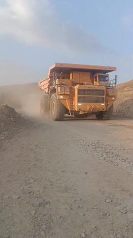 This media file captures a massive, bright yellow mining truck, identified as a BELAZ model with the number "112" prominently displayed. The truck is the primary subject, dominating the frame as it moves along a dirt road, kicking up a cloud of dust. The scene suggests an active mining operation in Almalyk, Uzbekistan, given the arid, dusty environment and the presence of such heavy machinery.

The time of day appears to be late afternoon or early evening, indicated by the warm, soft light and the long shadows. The weather seems to be clear and dry, conducive to the dusty conditions. The setting is a mine site, characterized by loose gravel, piles of earth or ore, and the vastness implied by the truck's scale. There are no people visible in the frame, emphasizing the immense size and power of the machine itself.

Notable details include the truck's enormous tires, the sturdy construction of its chassis and bed, and the detailed grille on the front. The text "YOSHIN" is visible on a label near the top right of the truck's cabin. The overall impression is one of industrial might and relentless operation. The activity is straightforward: the truck is in motion, likely transporting materials within the mine. The emotion conveyed is one of raw power and the scale of industrial endeavors.