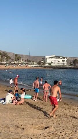 The video shows a crowded beach on a sunny day. The sand is light brown and there are many people in swimwear scattered across it, some sitting, some standing, and some walking. In the foreground, a group of people are sitting on blankets. Further back, people are interacting with each other, walking towards and away from the water, and some are in the ocean. The ocean is blue and calm, with small waves reaching the shore. In the background, there are palm trees, a white building with a modern design, and a mountain range with a clear blue sky above. There are also beach umbrellas and other beach accessories visible. A construction crane is visible in the distance behind the building.