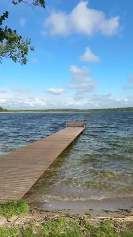 The video shows a wooden pier extending into a lake. The water is choppy and clear enough to see the lake bed near the shore. A small boat is docked beside the pier in some shots. The shoreline is grassy and wooded. The sky is blue with scattered clouds.