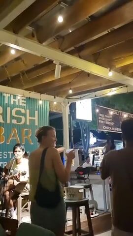 The video depicts an open-air bar at night, with an Irish bar sign visible in the background. A young woman is singing into a microphone, with a guitarist seated to her left. Another woman is seen from the back, partially blocking the view, and a man is standing to the right, recording the performance with his phone. A white bucket is placed on a stool in front of the singer, possibly for tips. String lights are hung across the wooden ceiling structure, illuminating the scene. A sign promoting free shots and shirts is also visible.