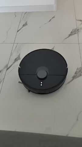 A robot vacuum cleaner is shown from above, moving across a tiled floor. The vacuum is dark gray and circular, with a prominent circular sensor on top. Two small lights are visible on the front of the vacuum. The floor is tiled with a white marble pattern. The vacuum moves from the upper right of the frame towards the bottom left, turning slightly as it moves.