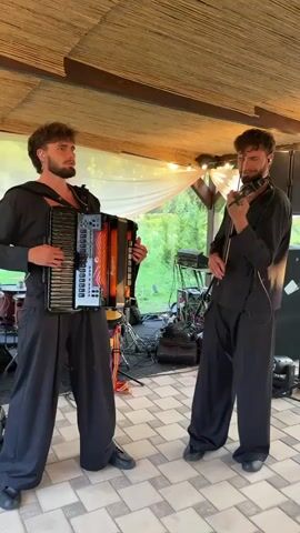 Two young men in black clothing are performing music outdoors under a bamboo-thatched roof. The man on the left is playing a large accordion, while the man on the right is playing a violin. Both men have dark, curly hair and beards. In the background, there are musical instruments, including drums, and stage lighting equipment. The ground is paved with square tiles. The overall atmosphere suggests a live musical performance.