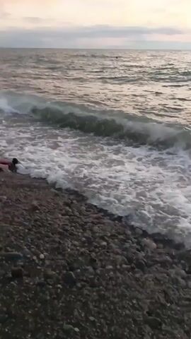 The video shows a person lying on a rocky beach near the water's edge. Waves are washing over them. The person appears to be wearing dark clothing and their feet are visible. The sky is overcast and the sea is choppy. The camera is positioned low to the ground, looking towards the water.