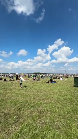 The video shows a wide shot of a grassy field on a sunny day with a bright blue sky filled with white, fluffy clouds. Many people are scattered across the field, sitting on blankets or chairs, or lying down. Some are interacting with each other, while others appear to be observing something happening off-camera. In the foreground, there is a large green dumpster. In the mid-ground, there are several clusters of people, some with umbrellas. Further in the distance, the field extends to a treeline with more people visible, suggesting a large event or gathering. The overall atmosphere appears relaxed and festive, likely a daytime outdoor event.