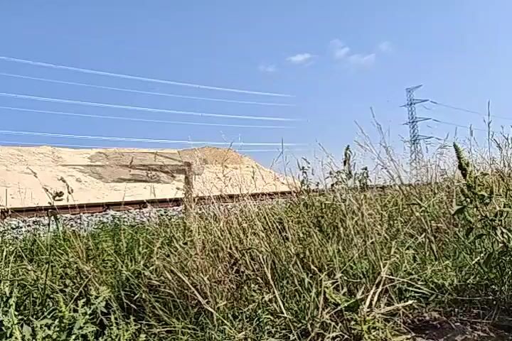 The video opens with a low-angle shot looking up at a clear blue sky with several white contrails stretching across it. In the middle ground, a large pile of sand is visible, likely a construction or industrial site, with a railroad track running in front of it. Tall, dry grass dominates the foreground, obscuring the lower portion of the frame. To the right of the sand pile, a tall electrical pylon stands against the sky. The scene appears to be outdoors on a sunny day. The video is static, with no discernible movement within the frame.