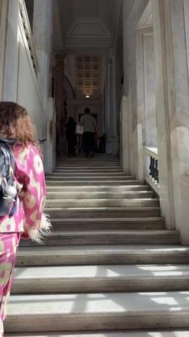 A person with long brown hair, wearing a pink and yellow patterned jumpsuit and a blue backpack, stands with their back to the camera at the bottom of a wide, grand marble staircase. The staircase is flanked by tall marble columns and walls, ascending into a large hall with an ornate, coffered ceiling. Several other individuals are visible ascending the stairs further in the distance; one person in a light grey shirt and dark pants is walking up, another in dark clothing also walks up, and two more are near the top of the stairs, one standing and one seated on the steps. The foreground person remains stationary throughout the video.