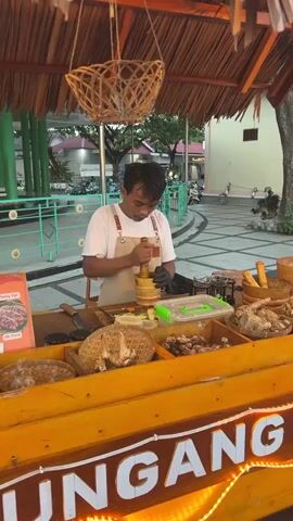 A man wearing a white t-shirt and an apron is standing behind a wooden stall and grinding ingredients in a mortar with a pestle. The stall has a thatched roof and is decorated with string lights. In front of the man, there are various ingredients in bowls, including what appears to be ginger and garlic. A sign with text in a foreign language is visible on the left. The background shows a paved area with some trees and buildings. The man is wearing black gloves.
