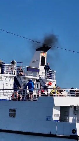 The video shows the stern of a white ferryboat sailing under a clear blue sky. The ferry is crowded with passengers on its upper decks. A prominent white smokestack emits a thick plume of black smoke into the air. Several passengers are visible on the open decks, some seated, others standing. A person in a blue jacket stands near the railing of the upper deck, facing away from the camera. Another person in a blue shirt stands on a higher platform near the smokestack, appearing to look out at the sea. A red flag is visible on the right side of the upper deck. The name of the port and registration number are partially visible on the hull of the ship at the bottom right of the frame.