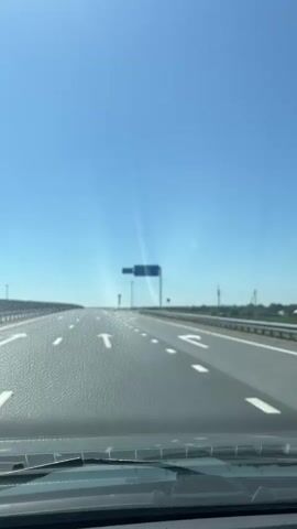 The video captures a first-person perspective from inside a vehicle traveling on a highway under a clear blue sky. The road is paved and has white lane markings, with arrows indicating directions. In the distance, a blue road sign is visible. Text on the sign reads "МОСКВА 578", "НИКОЛЬСКОЕ 0.2", and "ДОЛИНА 2". There are guardrails along the side of the road and some vegetation and distant buildings can be seen. A traffic sign with a red and white chevron pattern is also visible. The bright sky and sunlight suggest it is daytime, likely a clear and warm day. The overall scene depicts a typical highway journey through a rural or semi-rural landscape.