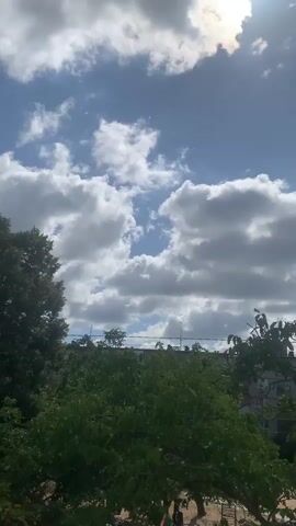 The video shows a time-lapse of a sky with clouds and some trees in the foreground. The sun is visible in the upper right corner, partially obscured by clouds. The clouds are white and fluffy, with some darker areas suggesting depth and shadow. The sky is blue. The trees are green and lush, suggesting a warm season. There are some buildings visible in the background, peeking out from behind the trees. The overall scene is peaceful and serene.