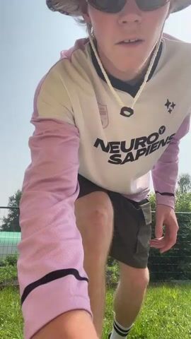 A person wearing a bucket hat, sunglasses, a long-sleeved shirt with pink sleeves and the text NEURO SAPIENS in black, and cargo shorts is performing a low to the ground stunt on a grassy area. The person begins by standing and then bends down and throws their legs up in the air. In the background, there is a fence, some greenery, and a bright sky.
