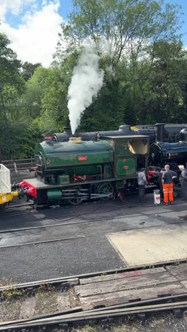 A vintage green steam locomotive with white steam emitting from its stack is stationary on train tracks.  Several individuals are gathered around the locomotive, some appearing to be performing maintenance or cleaning.  One person is wearing orange overalls.  Behind the green locomotive, another black steam locomotive is visible. The scene is outdoors, with trees and foliage in the background, under a cloudy sky.  Wooden train tracks are in the foreground.