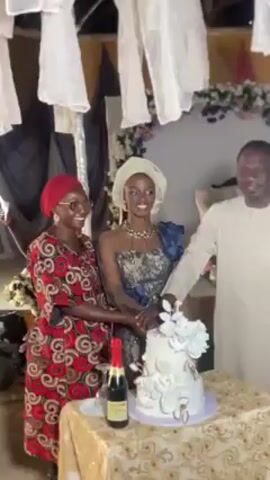 Here's a description of the video:

The video shows three people – a bride standing between a woman and a man – cutting a wedding cake. The bride is wearing a light-colored head wrap, and a detailed blue and gray dress. The woman to her left is wearing a red head wrap and a patterned red dress. The man to her right is wearing a light-colored shirt. They are all standing behind a table covered with a gold patterned cloth. On the table, there's a white three-tiered cake decorated with white flowers, and a bottle of champagne.  The backdrop behind them is decorated with white fabric and flowers.