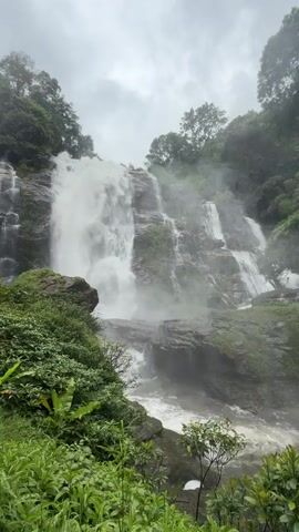 A wide shot captures a multi-tiered waterfall cascading down a rocky, forested mountainside. The water is white and frothy, creating a mist that hangs in the air, obscuring the upper portions of the falls and the surrounding trees. Lush green foliage, including large-leafed plants and dense undergrowth, fills the foreground and frames the left side of the waterfall. The sky above is overcast and grey, suggesting a cloudy or rainy day. The scene is entirely natural, with no visible human presence or man-made structures. The video appears to be a static shot, allowing the viewer to observe the continuous flow of the waterfall and the atmospheric conditions.