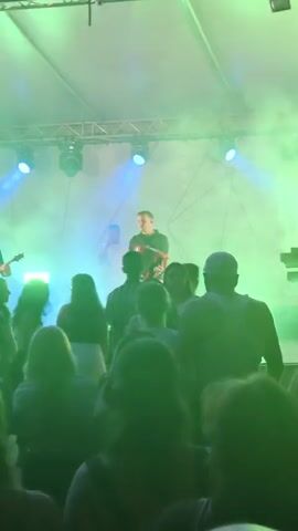 The video shows an outdoor concert stage bathed in green and blue stage lighting. In the foreground, the backs of a crowd of people are visible, their faces obscured by shadow and the angle of the shot. On stage, three musicians are performing. The musician on the far left is playing a guitar and wearing a black t-shirt. To his right, a man in a white t-shirt is partially visible. The central figure is a man in a black t-shirt and light-colored pants, actively playing a red accordion. The accordion has "Roland" printed on it. The stage has a white backdrop and a lighting rig above. The overall atmosphere appears to be lively and energetic.