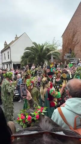 The video captures a lively street parade, likely a celebration or festival. A large crowd of onlookers lines the street, many holding up their phones to record the procession. Performers dressed in elaborate green costumes with leaf-like fringes and floral crowns walk down the street, some carrying musical instruments and swords. A large drum with a painted green face is prominently featured. The environment includes buildings of various colors and architectural styles, a palm tree, and street signs. The overall atmosphere is festive and celebratory.