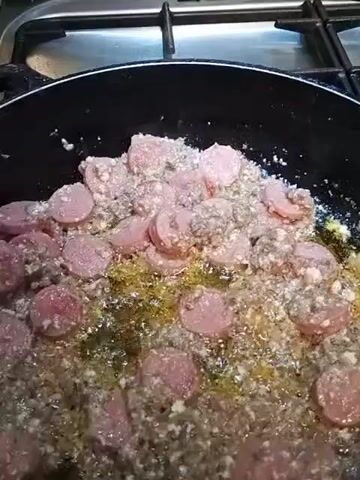 A close-up, overhead shot shows sliced sausages and ground meat cooking in a black frying pan on a stovetop. The pan is filled with oil, and the food is sizzling and browning. The stovetop has a metallic grate and knobs, suggesting a kitchen environment. The lighting is dim, with the primary illumination coming from the cooking process itself. The video appears to be a short, unedited clip with no discernible narrative or specific action beyond the cooking.